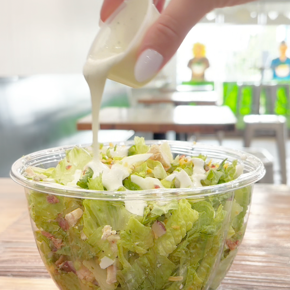 Hand pours creamy dressing over a fresh chopped salad in a clear bowl on a wooden restaurant table.